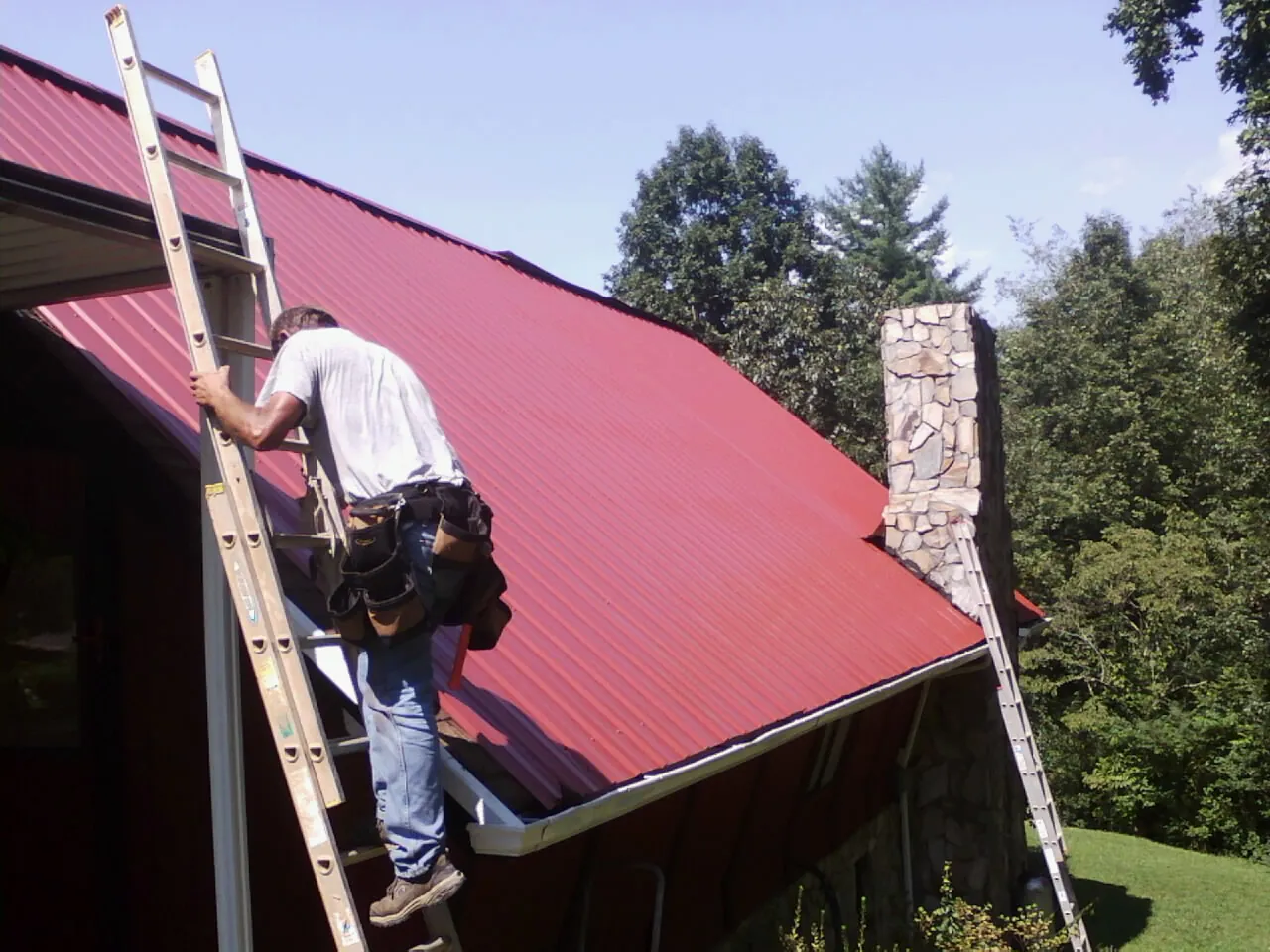 Roofer on a ladder beside a red metal roof and stone chimney — Soffit & Fascia Repair by Roofing Brothers in Morrison